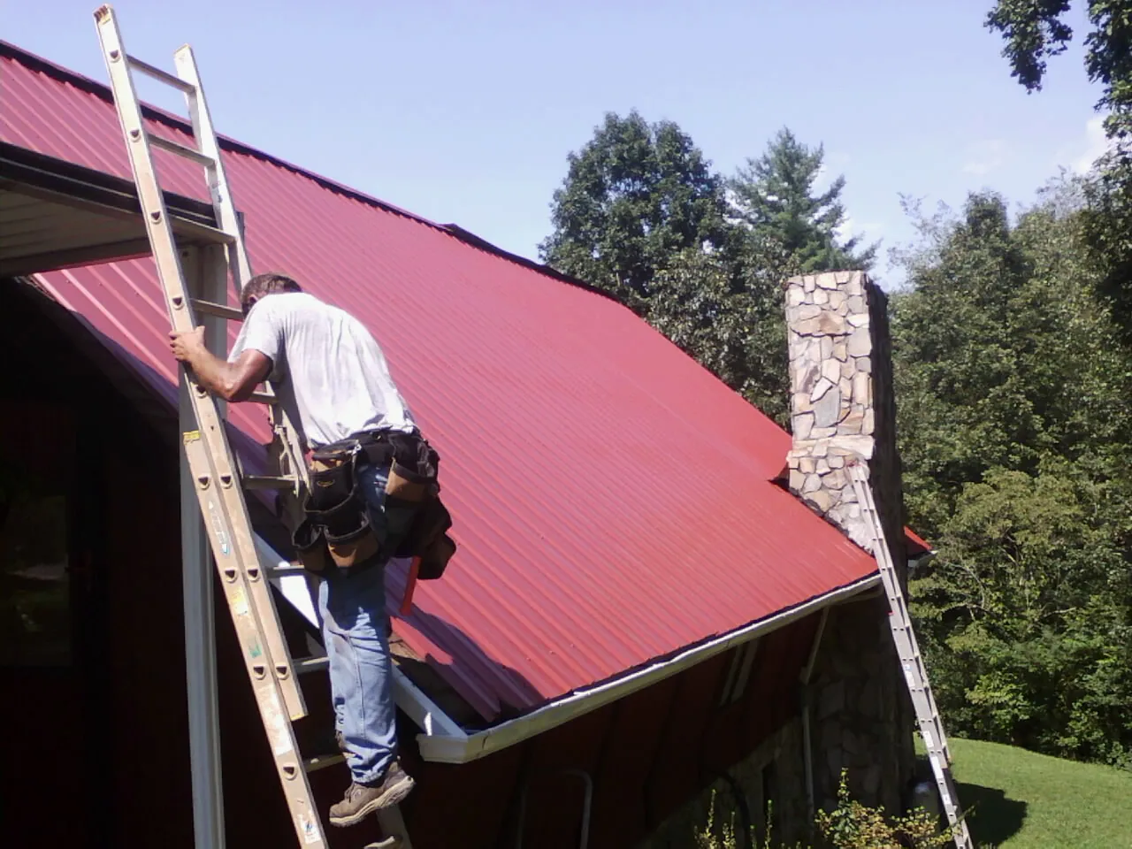 Roofer on a ladder beside a red metal roof and stone chimney — Soffit & Fascia Repair by Roofing Brothers in Meadowview